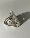 A textured sterling silver ring shaped like a stylized wing or leaf, photographed on a plain white surface with a sharp shadow.