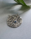 Silver textured round pendant with a blu sapphire on a light gray background with a green leaf