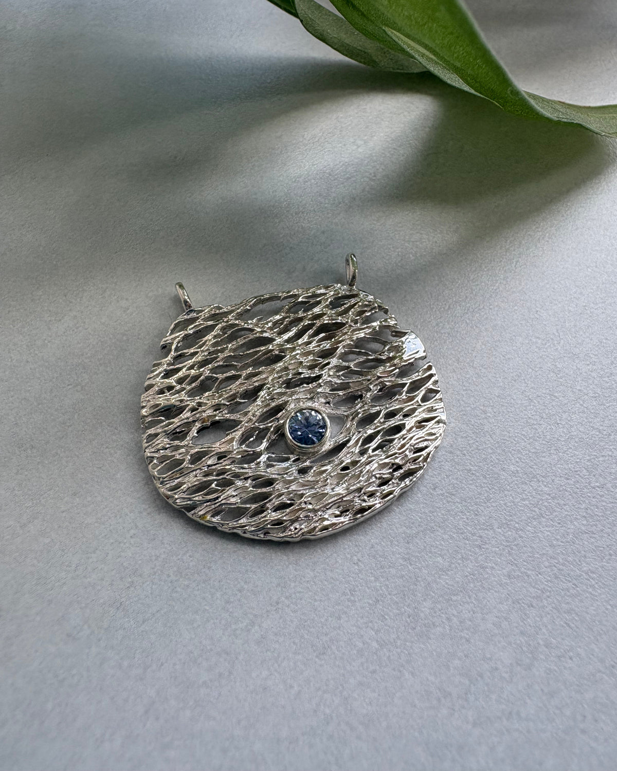 Silver textured round pendant with a blu sapphire on a light gray background with a green leaf