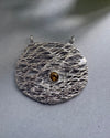 Silver textured brooch with a central gemstone on a gray background