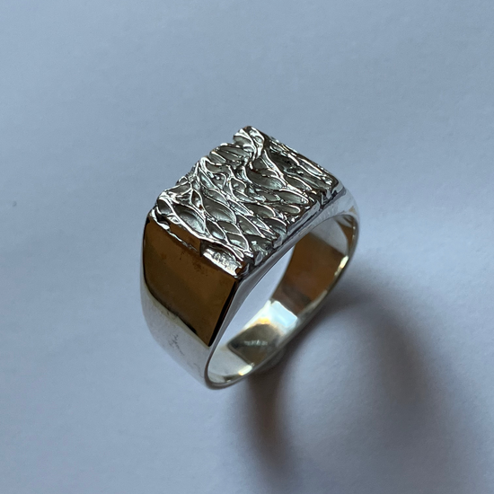 A silver signet ring with a heavily textured, hammered top and polished band, resting on a light gray surface.