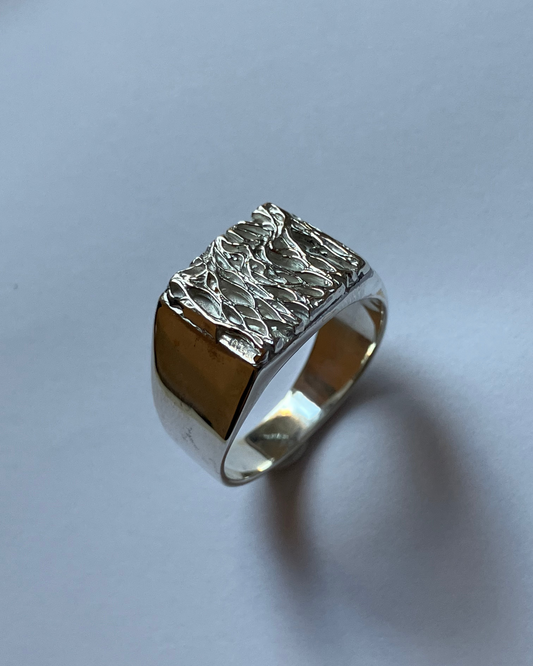 A silver signet ring with a heavily textured, hammered top and polished band, resting on a light gray surface.