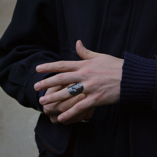 A close-up of a person's hands wearing a chunky silver ring with a textured, organic pattern, styled with a dark navy jacket and ribbed navy sweater.