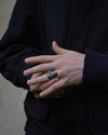A close-up of a person's hands wearing a chunky silver ring with a textured, organic pattern, styled with a dark navy jacket and ribbed navy sweater.