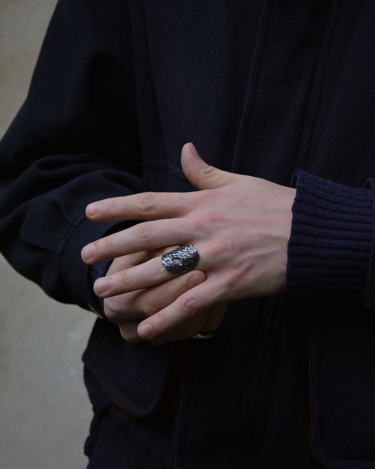 A close-up of a person's hands wearing a chunky silver ring with a textured, organic pattern, styled with a dark navy jacket and ribbed navy sweater.
