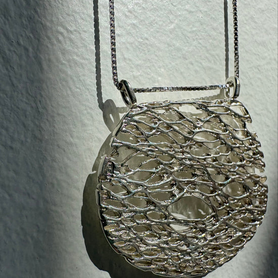 A silver-toned oval pendant necklace with an intricate, openwork web-like mesh pattern, hanging against a textured off-white wall with dramatic side lighting.