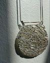 A silver-toned oval pendant necklace with an intricate, openwork web-like mesh pattern, hanging against a textured off-white wall with dramatic side lighting.