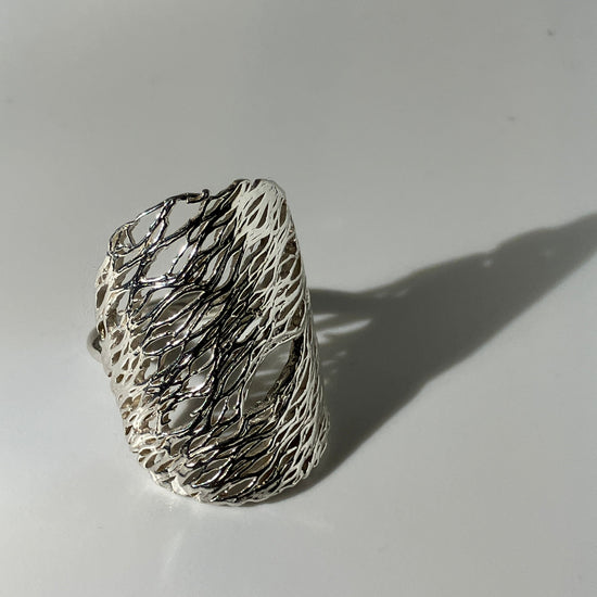 A wide silver ring with a textured, organic wire-like pattern, photographed on a light gray surface with a soft shadow.