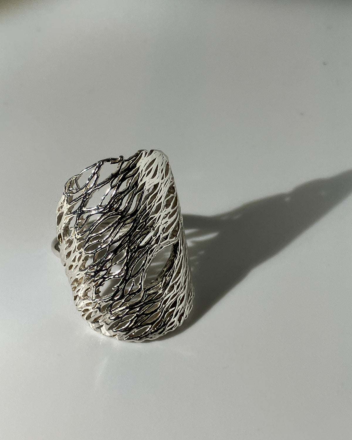 A wide silver ring with a textured, organic wire-like pattern, photographed on a light gray surface with a soft shadow.