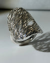 A silver ring with a highly textured, organic, tree-like branching structure, photographed on a white surface with a soft shadow cast to the right.