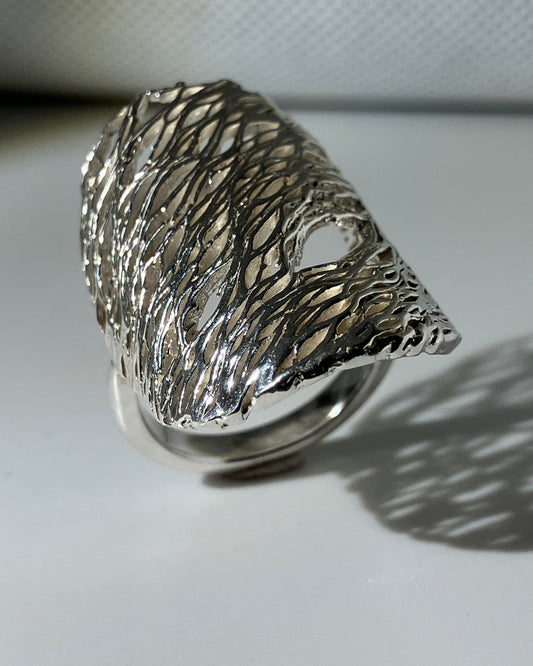 A silver ring with a highly textured, organic, tree-like branching structure, photographed on a white surface with a soft shadow cast to the right.