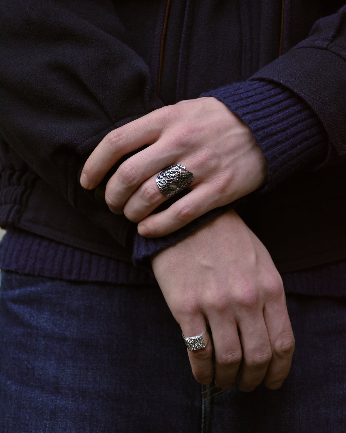 A close-up of a person wearing a dark navy jacket and blue jeans, showcasing two silver rings: a large textured band ring on the middle finger and a smaller patterned ring on the ring finger.