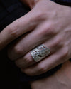 A close-up of a hand wearing a wide, textured silver ring with an organic, tree-like openwork pattern against a dark background.