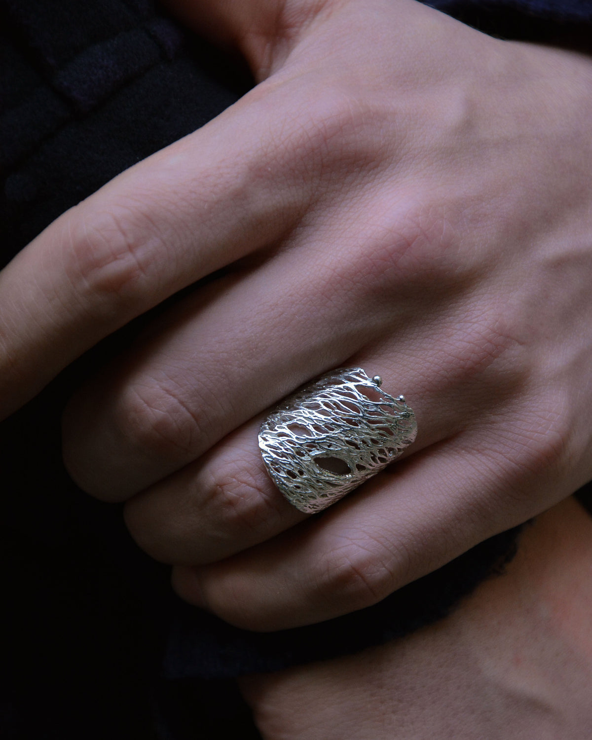 A close-up of a hand wearing a wide, textured silver ring with an organic, tree-like openwork pattern against a dark background.