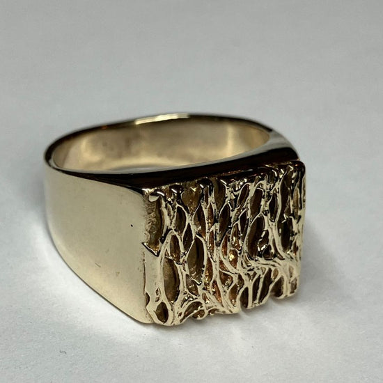 A gold signet ring with a textured, organic patterned face, photographed on a neutral light gray background.