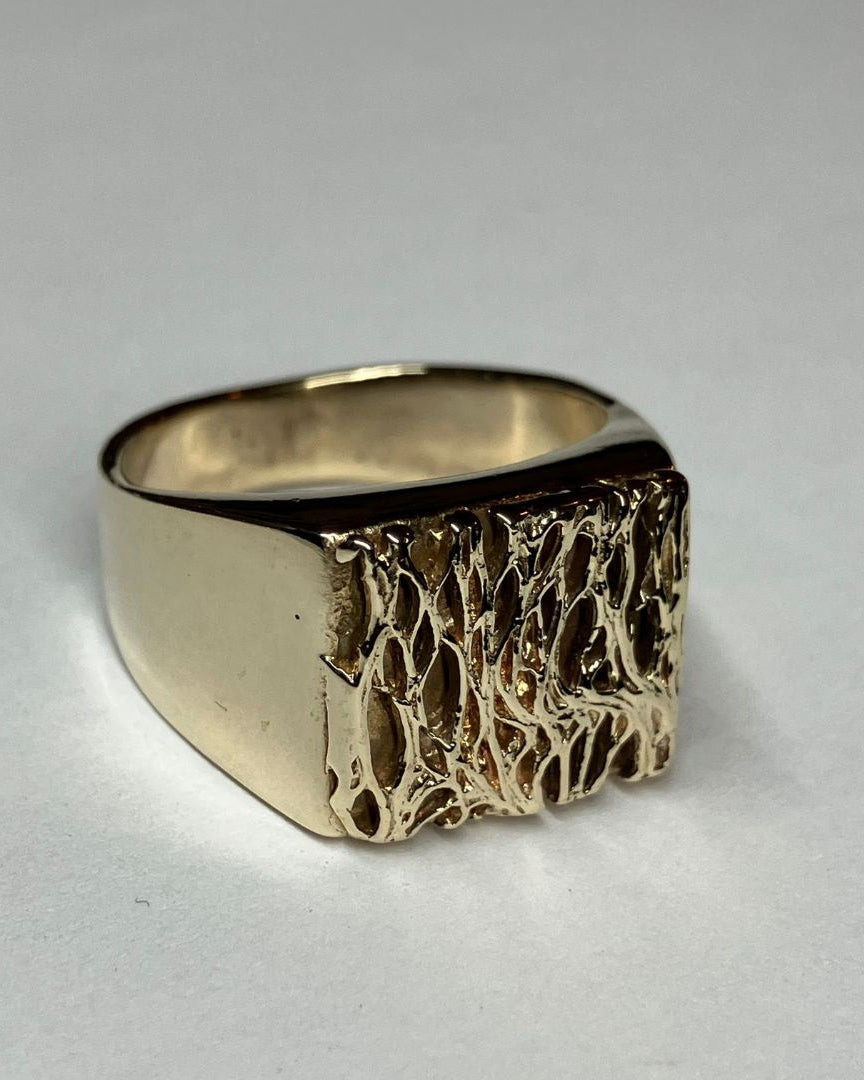 A gold signet ring with a textured, organic patterned face, photographed on a neutral light gray background.