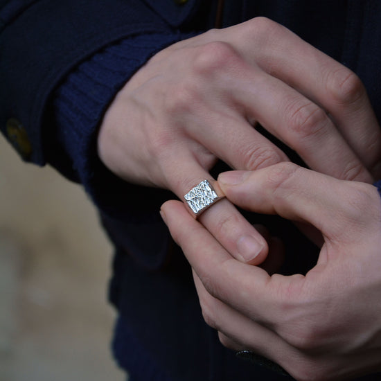 A close-up of a person's hands wearing a silver square ring with a textured surface on the finger, wearing a dark navy blue sweater.
