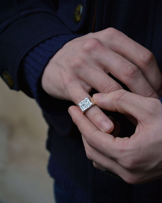 A close-up of a person's hands wearing a silver square ring with a textured surface on the finger, wearing a dark navy blue sweater.