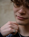 A close-up lifestyle shot of a person wearing a chunky silver ring with a textured top paired with a silver chain necklace and a dark patterned sweater.