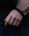 A close-up of a person's hand wearing a chunky, textured silver ring, with a second silver ring on the wrist, set against a dark navy sweater and jeans.