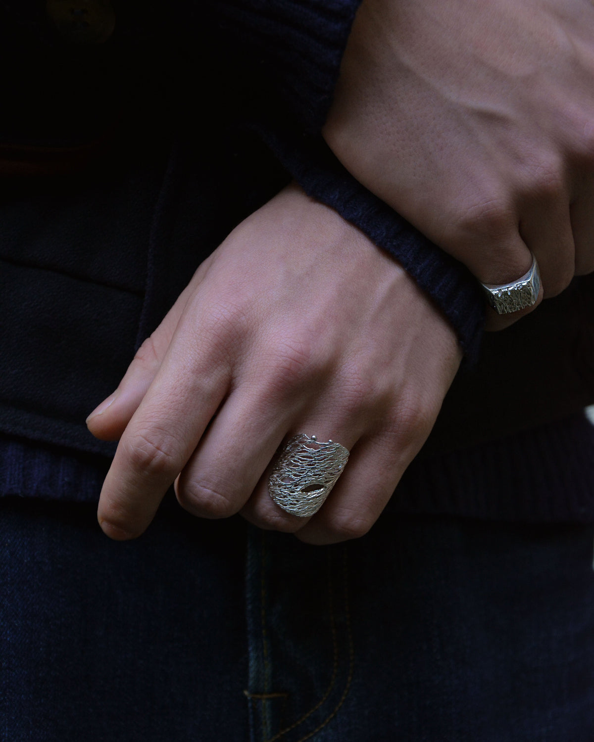 A close-up of a person's hand wearing a chunky, textured silver ring, with a second silver ring on the wrist, set against a dark navy sweater and jeans.