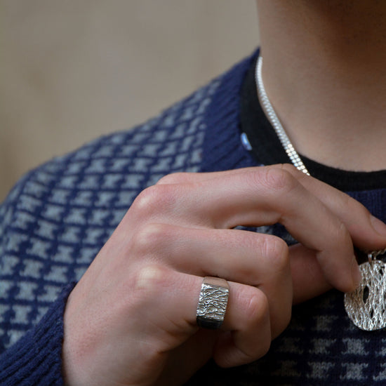 A close-up lifestyle shot of a man wearing a textured silver pendant necklace and a matching textured silver ring on his finger, wearing a navy and white patterned sweater.