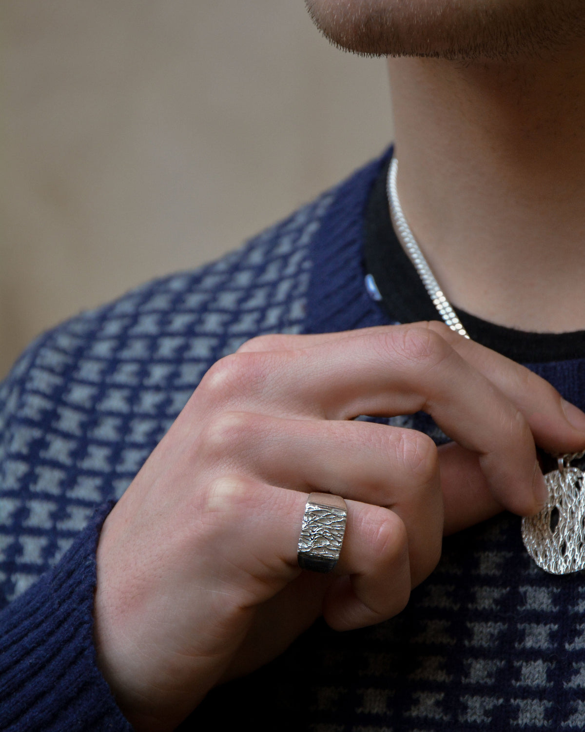 A close-up lifestyle shot of a man wearing a textured silver pendant necklace and a matching textured silver ring on his finger, wearing a navy and white patterned sweater.