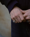 A close-up lifestyle shot of a person's hands clasped together, wearing a silver ring with a textured, set against a dark navy sweater and a soft beige background.