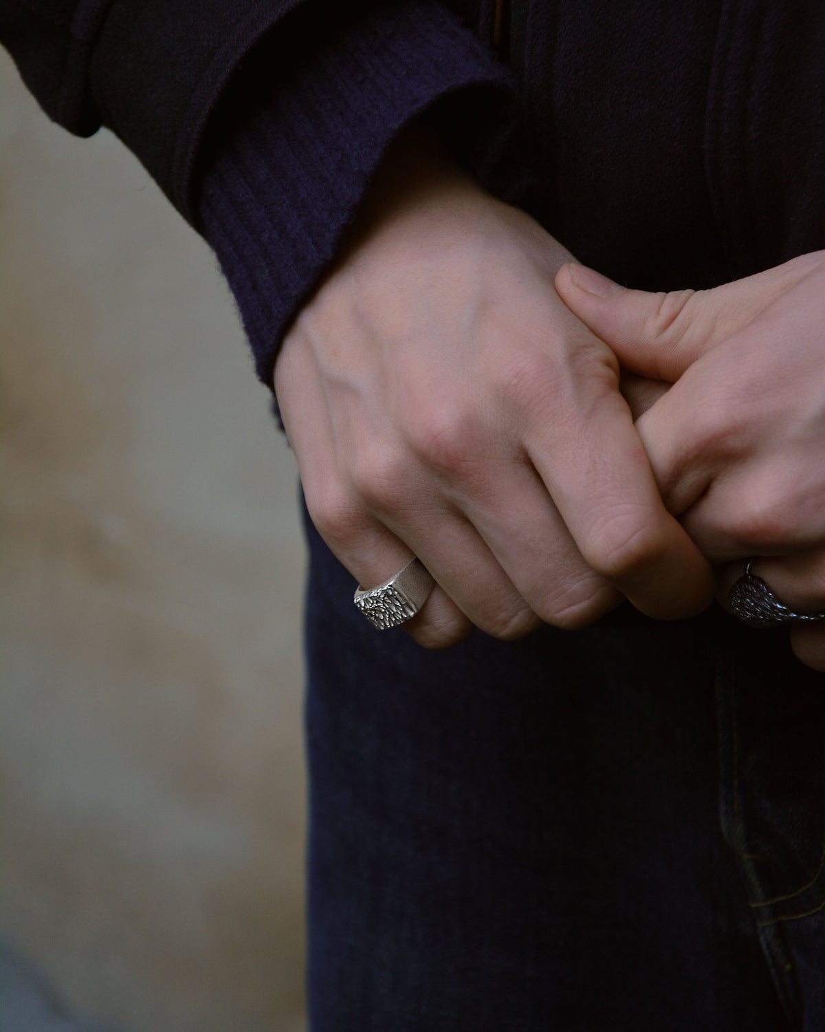 A close-up lifestyle shot of a person's hands clasped together, wearing a silver ring with a textured, set against a dark navy sweater and a soft beige background.