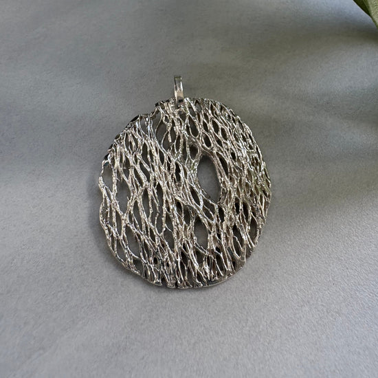 Silver circular lace-like pendant on a light gray surface with a leaf in the corner.