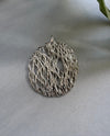 Silver circular lace-like pendant on a light gray surface with a leaf in the corner.