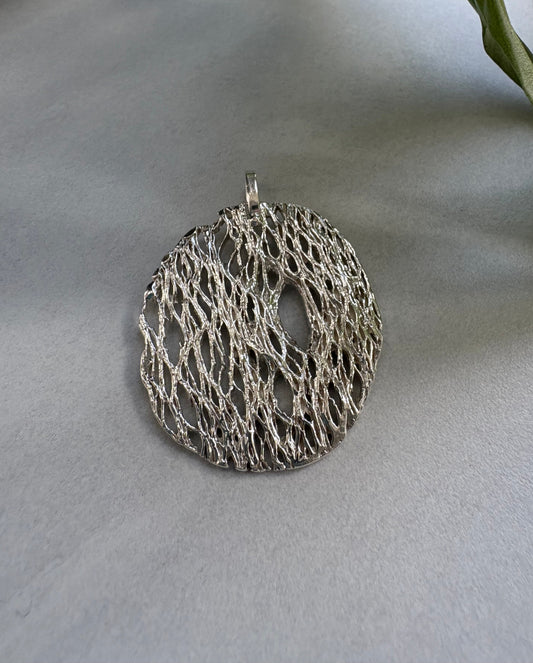 Silver circular lace-like pendant on a light gray surface with a leaf in the corner.