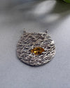Silver textured  pendant with a yellow gemstone on a gray surface
