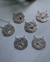 Silver circular metal textured pendants with different coloured gemstones on a gray background