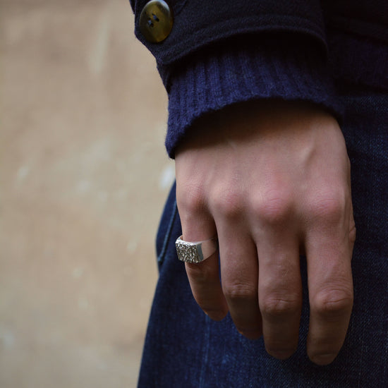A close-up shot of a person's hand wearing a textured silver signet ring, dressed in a dark navy blue knit sweater and matching navy blue denim skirt, with a neutral beige wall in the background.