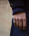 A close-up shot of a person's hand wearing a textured silver signet ring, dressed in a dark navy blue knit sweater and matching navy blue denim skirt, with a neutral beige wall in the background.