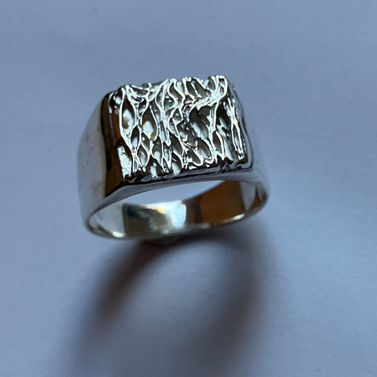 A silver signet ring with a heavily textured, hammered top surface, photographed on a plain light gray background.