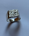 A silver signet ring with a heavily textured, hammered top surface, photographed on a plain light gray background.