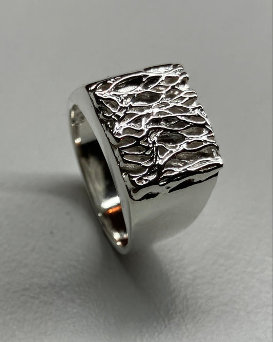 A silver ring with a square, heavily textured and hammered top, photographed on a neutral gray surface.
