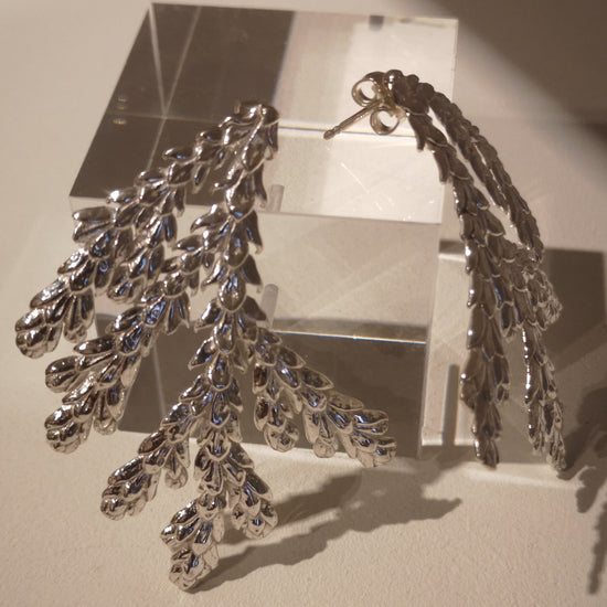 A pair of silver-toned dangle earrings with an intricate, leafy or coral-like textured design, displayed on a clear acrylic block against a warm beige background.