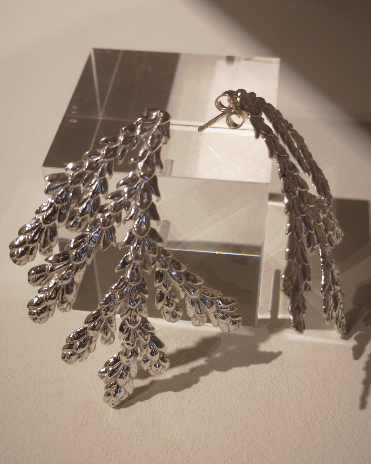A pair of silver-toned dangle earrings with an intricate, leafy or coral-like textured design, displayed on a clear acrylic block against a warm beige background.