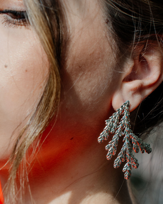 A close-up of a person's ear wearing a textured, silver-toned dangle earring with a leaf or pine-branch-like organic shape.