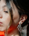A close-up lifestyle shot of a woman wearing a vibrant orange top and a sparkling silver leaf-shaped earring, set against a soft, blurred green background.