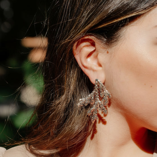 A close-up lifestyle shot of a woman wearing a pair of large, leaf shaped statement earrings.