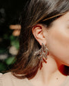 A close-up lifestyle shot of a woman wearing a pair of large, leaf shaped statement earrings.
