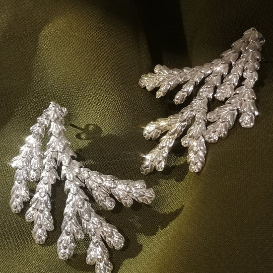 A pair of sterling silver leaf-shaped earrings displayed on a dark olive green fabric background.