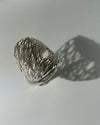 A wide silver ring with a textured, openwork wire-like design, photographed on a light gray surface with a prominent shadow.