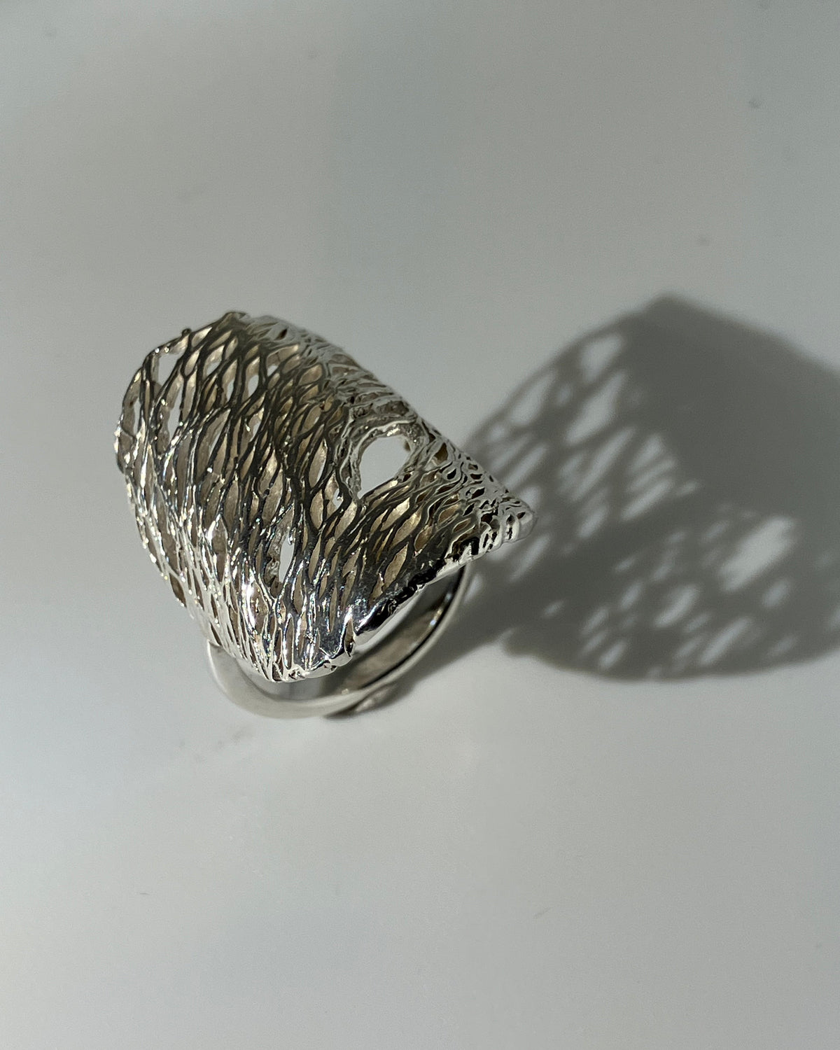 A wide silver ring with a textured, openwork wire-like design, photographed on a light gray surface with a prominent shadow.