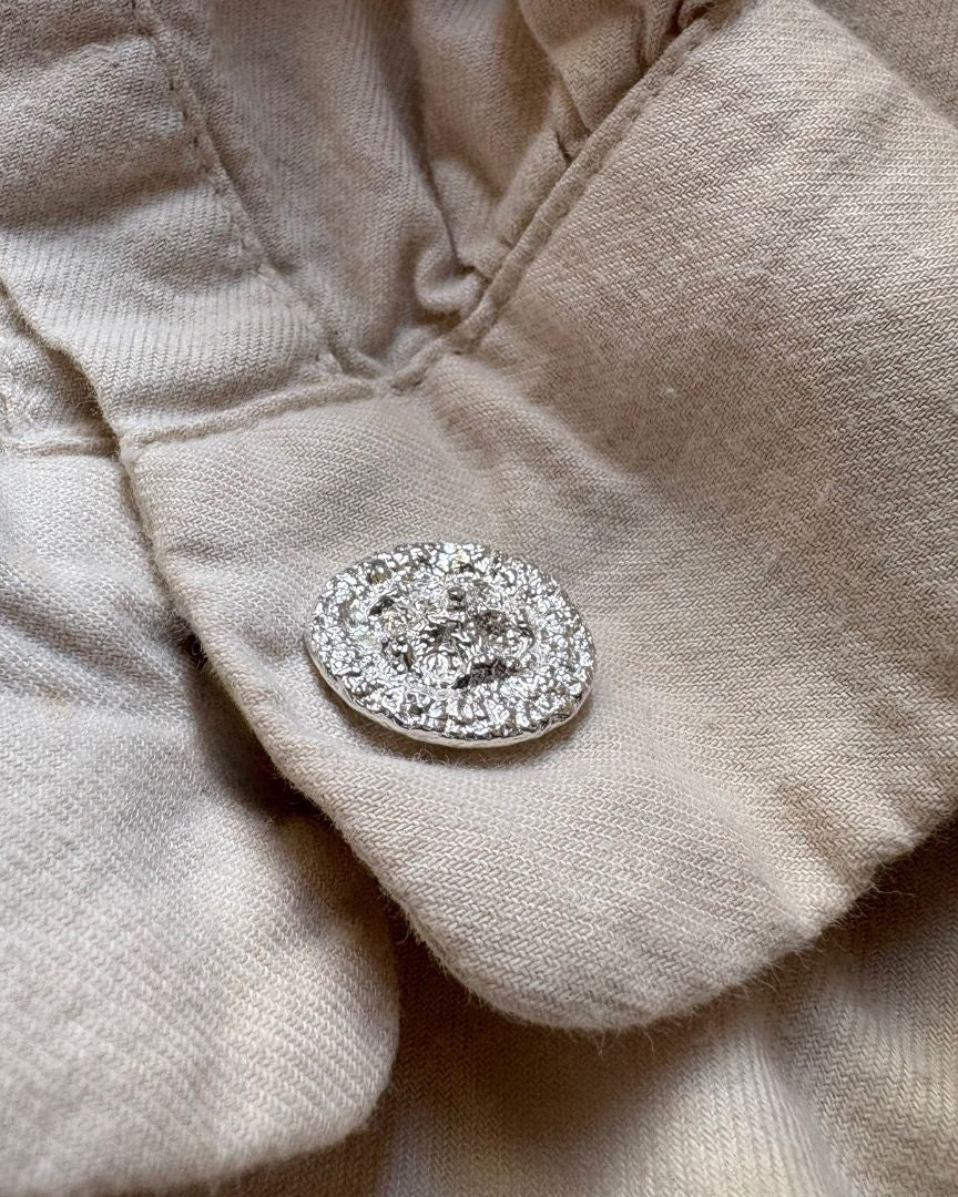 A close-up of a textured beige fabric garment featuring a single round silver cufflink set against a matching beige background.