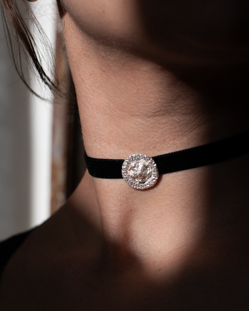 A close-up of a person's neck wearing a black velvet choker with a central silver-toned circular medallion embellished with intricate texture